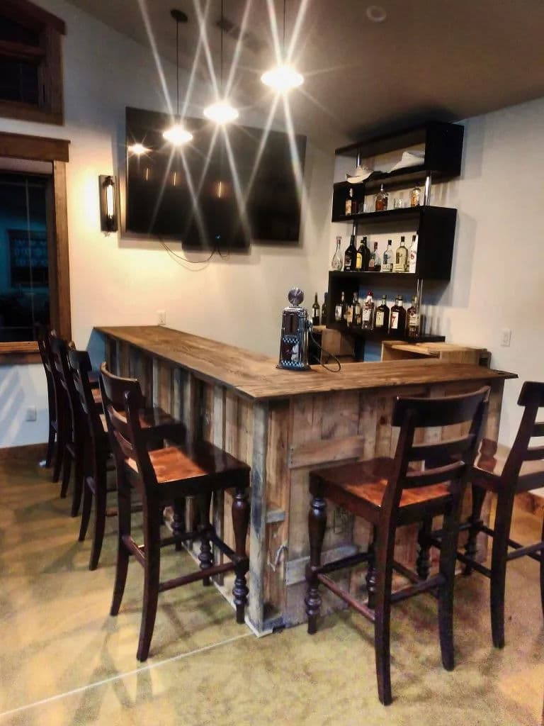 Modern home bar with wooden counter, stools, wall-mounted TV, and shelves of liquor.