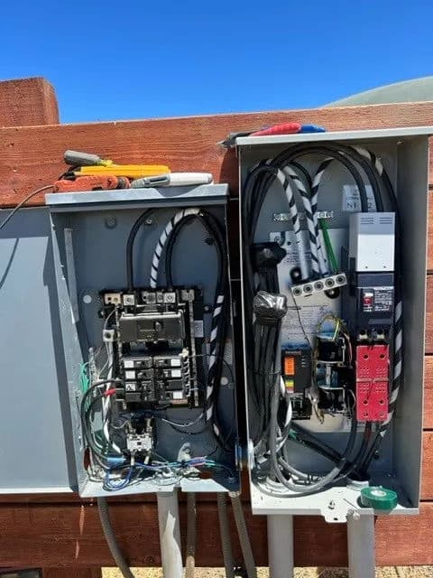 Electrical service panel with circuit breakers and wiring, mounted on a wooden fence.